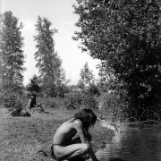 Antoine Ninamee and Loma Charlemain, two Native American boys on the Flathead Indian Reservation in western Montana, sit near a creek. The young man in the foreground wears a loincloth and squats down next to the creek scooping water into his hands. The other young man sits in the left background near a pile of clothing. He wears his hair in braids. Numerous trees stand along the creek.