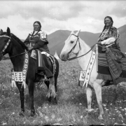 Two Native American women on the Flathead Indian Reservation in western Montana pose on horseback in a field. Both have long hair in braids; each wears a dress adorned with beads. The bridles on the horses are similarly adorned with beadwork. The women sit on patterned, wool blankets that cover their wood-frame saddles. Mountains aren the distance.