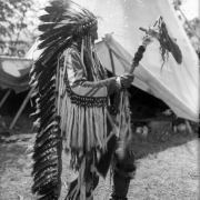 Joe Lamoose, a Native American on the Flathead Indian Reservation in western Montana, dances in front of a teepee on the reservation. He holds a pipe tomahawk adorned with fur and feathers and wears a long, feather headdress. Long, ermine fur tassles and beadwork adorn the sleeves of his buckskin shirt. He also wears beaded moccasins.