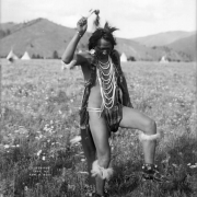 Joe Vanderburg (also knwn as Yellow Rock), a Native American man on the Flathead Indian Reservation in western Montana, dances in a field on the reservation. He wears a striped loincloth and several long, beaded necklaces. Strips of fur and bells are tied around his lower legs and his arms. Additional straps of fur are wrapped around his long braids. Several teepees are in the distance at the base of a ridge of mountains.