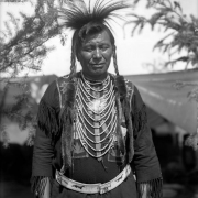 Loma Standing Bear, a Native American man on the Flathead Indian Reservation in western Montana, poses for an informal, half-length portrait. He wears his long hair in braids with strips of fur wrapped around their ends. He also wears a breastplate, a belt with beadwork depicting horses, a beaded-flower shirt, and tassels on the sleeves and bottom of his shirt.