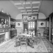 Interior view of a dining room in Denver, Colorado. Decor includes a table carved griffin legs, an Oriental rug, silver urns and vases, a stuffed bird, a crystal punch bowl, and a sideboard with a silver serving set. A ram's head is mounted above the door.