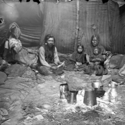A group of unidentified Native Americans sits playing cards inside a teepee on the Flathead Indian Reservation in western Montana. Numerous pillows and blankets are on the ground inside the teepee. A group of pots and containers is in the foreground.
