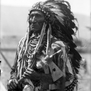 Joe Lamoose, a Native American man on the Flathead Indian Reservation in western Montana, poses for an informal, half-length portrait. He wears a feather headdress and holds a pipe tomahawk in his hands. He wears several beaded necklaces, ermine fur tassles and beads on the sleeves of his shirt, and a beaded pouch on his belt.