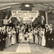 View of a wedding party at Lakeside Park in Lakeside (Jefferson County), Colorado. Well dressed men, women, boys and girls pose by signs and banners: "Ice Sitting Contest Starts 9 O'clock Tuesday Nite," and "Your Master of Ceremonies is Henry Polk." Some hold flower bouquets and shepherds' crooks. The bride and groom stand under a wicker arch.