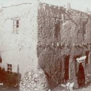 View of a dilapidated adobe house located at 215 E. De Vargas Street, Santa Fe (Santa Fe County), New Mexico. The house has vigas, chimneys and a stone buttress, and is thought to be the oldest house in the city. Shows a woman with a basket on her head and a dog.