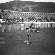View of a bullfight in a small stadium, in Gillett, Colorado, show two men in bullfighting attire. Two matadors hold capes, a third sits atop a bull who is ready to charge. The bullfighters are in a wooden corral, while observers watch from bleachers. The matador's costumes include hat, embroidered bolero jacket, epaulets & pants.