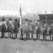 A group of Mexican vaqueros poses on horseback in a dirt arena for Buffalo Bill's Wild West Show at Ambrose Park in Brooklyn, New York. The men all wear sombreros and bolero jackets. A man in the center of the group holds a Mexican flag on a pole in his right hand.