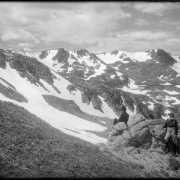 Looking to snow-covered Arapahoe Peaks, Colorado, from Denver and Salt Lake Railroad's Moffat Road; two men posed at boulders right foreground, one in suit and derby hat, and the other in knee-high laced boots, trousers tucked in boots, suspenders, long sleeved shirt with turtleneck and felt hat.