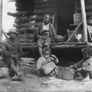 African American (Black) children and a man sit in front of a log cabin in an unknown location. The man wears a jacket, short pants, and sits on the porch near wood buckets and chairs. One boy sits on a pile of rocks that serve as steps. He wears a hat, wool jacket and torn pants. Two children use spoons to eat from a large iron pot. All of the children are barefoot.