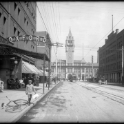 View down 17th (Seventeenth) street to Union Station prior to construction of the Welcome Arch; shows passengers boarding street railway car in center background, Oxford Hotel at left, Hendrie and Bolthoff Manufacturing and Supply Company at right, horse-drawn carriages on the street, and a young boy with knickers approaching bicycle left foreground; many men sit in front of the Oxford Hotel.