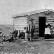 View of Grand Junction Town & Development Company's first office, on 2nd (Second) and Ute Streets, in Grand Junction, Colorado, with R.D. Mobley and Mrs. Jackson posed in front of the small one-room log cabin with sod roof. A "dress making" sign is attached to the cabin and a pile of firewood is between two posts supporting wire or string. Outhouses in back of the cabin show. The cabin is thought to be the first structure built in Grand Junction with R.D. Mobley the first postmaster.