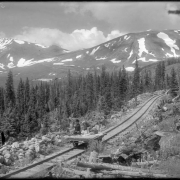 View of Argentine Central Railway narrow gauge track with push car near timberline, Argentine Pass, Clear Creek County, Colorado. Waldorf Mine distant center; Rocky Mountains with snowfields; man on push car with derby hat.