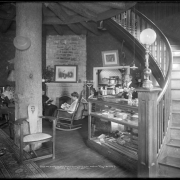 Hotel Glenisle, office area, Glenisle, Colorado, reached via Colorado and Southern Railway; interior view, man standing behind display case containing cigars, fishing poles and supplies with ledger on top; man and women reading magazines seated in rocking chairs in front of brick and stone fireplace; massive log center support, persian or oriental rug with spittoon; entryway into dining room behind display case.