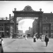 Welcome Arch at Union Station in Denver, Colorado; view of 17th (Seventeenth) street, street railway car number 248, men and women walking towards Union Station (not in view), horse-drawn carriages and a touring sightseeing coach parked at curb, Hendrie-Bolthoff building, Elk Hotel, bar, restaurant, drugstore, and the Struby-Estabrook Mercantile Company.