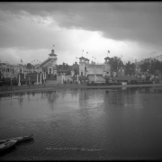View across Lake Rhoda towards the "White City" (later called Lakeside Amusement Park), Lakeside, Colorado, near Denver; shows the Big Splash ride, ferris wheel, Scenic Railway depot and two miniature steam locomotive engines and cars (train), roller coaster, clock, boats in lake, and rain clouds.