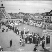 View of ride called the Chutes (or the Big Splash), "White City" (later called Lakeside Amusement Park), Lakeside, Colorado, near Denver; shows a large crowd of people gathered and watching water ride, edge of ferris wheel, other rides, a penny arcade, a large line by ticket gazebo, and wooden frame residences behind boundary of amusement park.