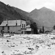 View east down mud and debris-filled street from the disastrous Cornet Creek flood on July 27, 1914, Telluride, Colorado; shows two-story false front, Iowa House on corner, other wood frame businesses, and residences. Men and woman make their way along streets on foot and horseback. San Juans and Ingram Falls are in background.