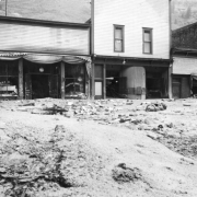 View north towards businesses on Colorado Avenue after the Cornet Creek flood on July 27, 1914, Telluride, Colorado. Mud and debris fill street in front of three false front wood frame structures between Sheridan Hotel and First National Bank, including the Phoenix Market.