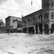 View northwest down Colorado Avenue after the Cornet Creek flood on July 27, 1914, Telluride, Colorado; shows First National Bank Building on corner of Fir and Colorado, three false front businesses, the Sheridan Hotel, the San Miguel County Courthouse, and mud and debris- filled street. Townspeople are near the bank and in distance in front of the Sheridan with a horse and automobile; electric and telephones lines cross street; chairs and tables are part of debris.