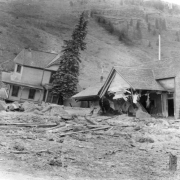 Oak Street filled with mud and debris from the disastrous Cornet Creek flood on July 27, 1914, Telluride, Colorado. Jack Hawkins' two-story wood frame Victorian with front wraparound porch and second-story bay window tilts on side from flood waters; smaller wood frame structure with front intersecting gables destroyed in foreground; cliffs in background.