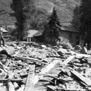 Two men stand on top of debris brought down by the Cornet Creek flood on July 27, 1914, Telluride, Colorado. Mud and debris fill street with damaged wood frame structures; Telluride Hospital is in background with building leased to San Miguel County Historical Society for use as a museum.