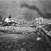 View south down Oak Street filled with mud and debris from the disastrous Cornet Creek flood on July 27, 1914, Telluride, Colorado. Townspeople survey damage. Jack Hawkins' two-story wood frame Victorian with front porch and second story bay window tilts on side from flood waters; smaller destroyed structure is next door; residences line street; church and back of San Miguel County Courthouse are in distance.