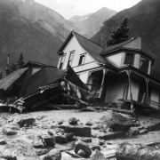 Exterior view of Jack Hawkins residence, on Oak Street, filled with mud and debris from the disastrous Cornet Creek flood on July 27, 1914, Telluride, Colorado; shows two-story wood frame Victorian with front porch and second story bay window tilting on side from flood waters, smaller destroyed structure, and the San Juans in background.
