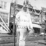 Close-up view of Chief of Midland Band, Fourth of July celebration, 1895, Victor, Colorado; shows man in costume with feathered headdress holding large pointed staff, a young boy in street-clothes with suspenders and another man in costume with feathered headdress standing on each side.  Background view is of commercial wooden frame false front buildings with boardwalk; a patriotic bunting hangs from cornice.
