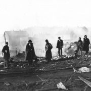 Six men survey destruction from second fire on April 29, 1896, Cripple Creek, Colorado; five men have sherriff badges, some wear overcoats, and one man wears knee-high rubber boots and carries a lunch bucket.