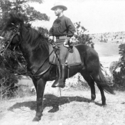 A cavalryman of the 6th Division of the U.S. Army poses on horseback in the foreground. He wears riding boots and gloves. Several trees are in the midground behind him, and a ridge is in the distance.