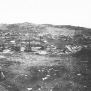 Destruction caused by second fire on April 29, 1896, Cripple Creek, Colorado; blocks of destroyed buildings in the commercial district, only foundations and twisted sheet metal remain, distant view of remaining buildings and residences.