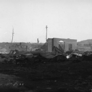 View of desolation caused by second fire on April 29, 1896, Cripple Creek, Colorado; stark remains, twisted sheet metal and debris cover the ground and smoke still rises from ashes; residence or building with hipped roof are in background.