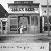 A saloonkeeper wearing an apron stands outside of a saloon in La Junta, Colorado in Otero County.  The nineteenth century, commercial storefront has bracketed cornices, two bays, storefront windows, a transom, and double entry doors.  Bottles of beer are visible in the window.  Signs: "Powell & Wonderly," "Zang's Beer," "Saloon."