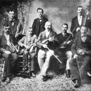 Reproduction of studio portrait of eight members of the La Junta Orchestra posing with their instruments. Three musicians stand, including the valve trombone player and the bass player.  Five musicians sit, including the trumpet player, the clarinet player, and the three violinists.  An oboe and a clarinet are at the feet of the clarinetist.