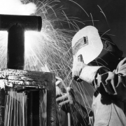 A student welds a metal frame with a threaded t-shaped pipe on the top of it in a welding class in Greeley, Colorado in Weld County.  He holds the welder in his right hand and wears a welding mask and heavy gloves that protect his hands and forearms.