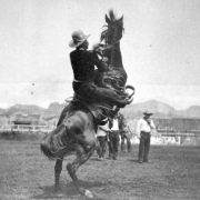 Antonio Esquivel, a champion Mexican vaquero and star of Buffalo Bill's Wild West Show, rides a bucking horse in a dirt arena during a performance of the show at Earl's Court in England. He wears a cowboy hat and a short, dark jacket. A group of cowboys stands in the background watching Esquivel ride. A backdrop with a landscape scene painted on it is at the far end of the arena.
