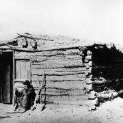 A miner sits outside a crudely built log cabin, Gunnison, Colorado, with his pick ax and shovel. The shack is known as the Sylvester Richardson cabin and the first one in town. The structure has logs of varying sizes chinked with mud, a low sagging front gable roof, hewn log front door and window opening.