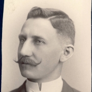 Studio bust portrait of Clark Hammond Rice, possibly during his senior year at Denver High School in Denver, Colorado. Rice's short hair is parted on the side and his bangs are styled in an upward wave. He has a handlebar moustache and he wears a morning coat with a silk tie and a shirt with an upright collar.