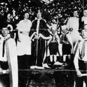 The Queen stands on stage with a crown and long robe at the 1923 Winter Sports Carnival Ball, Hot Sulphur Springs, Grand County, Colorado. Other participants and escorts stand around her. A pennant hangs in decorative evergreen backdrop: "$$ Winter Sport."