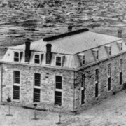 View of the Territorial Prison (later the Colorado State Penitentiary) building in Canon City (Fremont County), Colorado. Shows a two story stone building with a mansard roof, chimneys, and dormers.