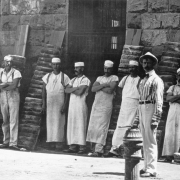 Prisoners in aprons and bakers caps pose near bread stacked against a stone wall of the bakery at the Colorado State Penitentiary in Canon City (Fremont County), Colorado. A man in a striped shirt, a bow tie, suspenders, light pants and a hat poses near the bakers. A fire hydrant is near the men.