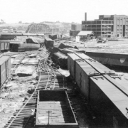 View of the Denver and Rio Grande Western Walker freight yards after the Arkansas River flood in Pueblo, Colorado. Shows twisted and damaged railroad tracks and overturned railroad freight cars. The Atchison, Topeka and Santa Fe bridge, the collapsed D. &.R.G.W. bridge, and the Nuckolls Packing plant are in the distance.