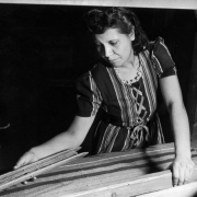 A Spanish American woman, a participant in a Federal Work Projects Administration program, holds a wool shuttle and the beater on an upright wooden loom as she weaves. She wears a striped dress with criss-crossed ribbon on the bodice, and a lace collar.