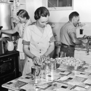 A man and two women work in a kitchen canning apricots or peaches for the hot lunch project for the Work Projects Administration in Colorado. One woman stands at a cast iron stove and sterilizes a can in hot water while the other woman stands at a kitchen table and packs fruit into a can. The man seals the can with a hand cranked machine attached to a counter. The table has a linoleum top decorated with an Art Deco pattern.
