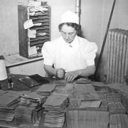 A woman in a white uniform and a white cap that reads: "First Aid," sits at a table and stamps paper sacks for surplus commodities at the State Grocery Warehouse at 1535 13th (Thirteenth) Street in Denver, Colorado. Piles of paper bags, an ink pad and a spool of string are on the table. A wooden file with cubbyholes is on a shelf. Labels attached to the file read: "Not To Be Sold" and "Graham Flour."
