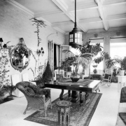 View of a sunroom in the Crawford Hill house at 969 Sherman Avenue, in the Capitol Hill neighborhood of Denver, Colorado.  The room has a tiled floor, brick walls, and a coffered ceiling. A large, square table with a pedestal and four carved legs is in the center of the room. Wicker and slipcovered furniture is scattered throughout the room. A large metal lantern hangs over the table. Shows potted plants , a mirror, and large windows with sheer blinds and shades. An oriental rug is under the table.