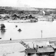 View of flood waters and railroad tracks in Pueblo, Colorado. A woman walks on a railroad embankment; houses, garages, out buildings and tracks are washed up against the embankment. Shows damaged houses and buildings, factories, and a bridge in the flood water and in the distance across the Arkansas River. The Nuckolls Packing Plant is in the distance.