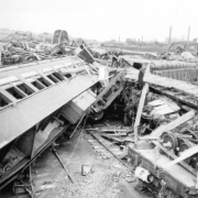 View of an over-turned railroad passenger car, box car and debris after a flood in Pueblo, Colorado. A man picks through the rubble. An intact Atchison, Topeka & Santa Fe bridge, a damaged Denver & Rio Grande Western Bridge, the Arkansas River, and the American Smelting and Refining Company smelter are in the distance.