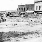 People walk through the debris outside stores destroyed by the Arkansas River flood, on Union Street in Pueblo, Colorado. Signs on commercial buildings read: "Weston's Cafe," "Cut Price Market," "Clothing" and "T. Flynn, Saddles and Harn[esses.]"